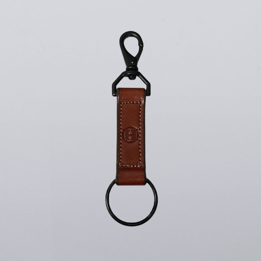 The Handy-Dandy Keychain by Hanks Belts features a brown leather strap with subtle stitching, embossed logo, black metal lobster clasp, and round keyring—crafted for classic style and everyday use.