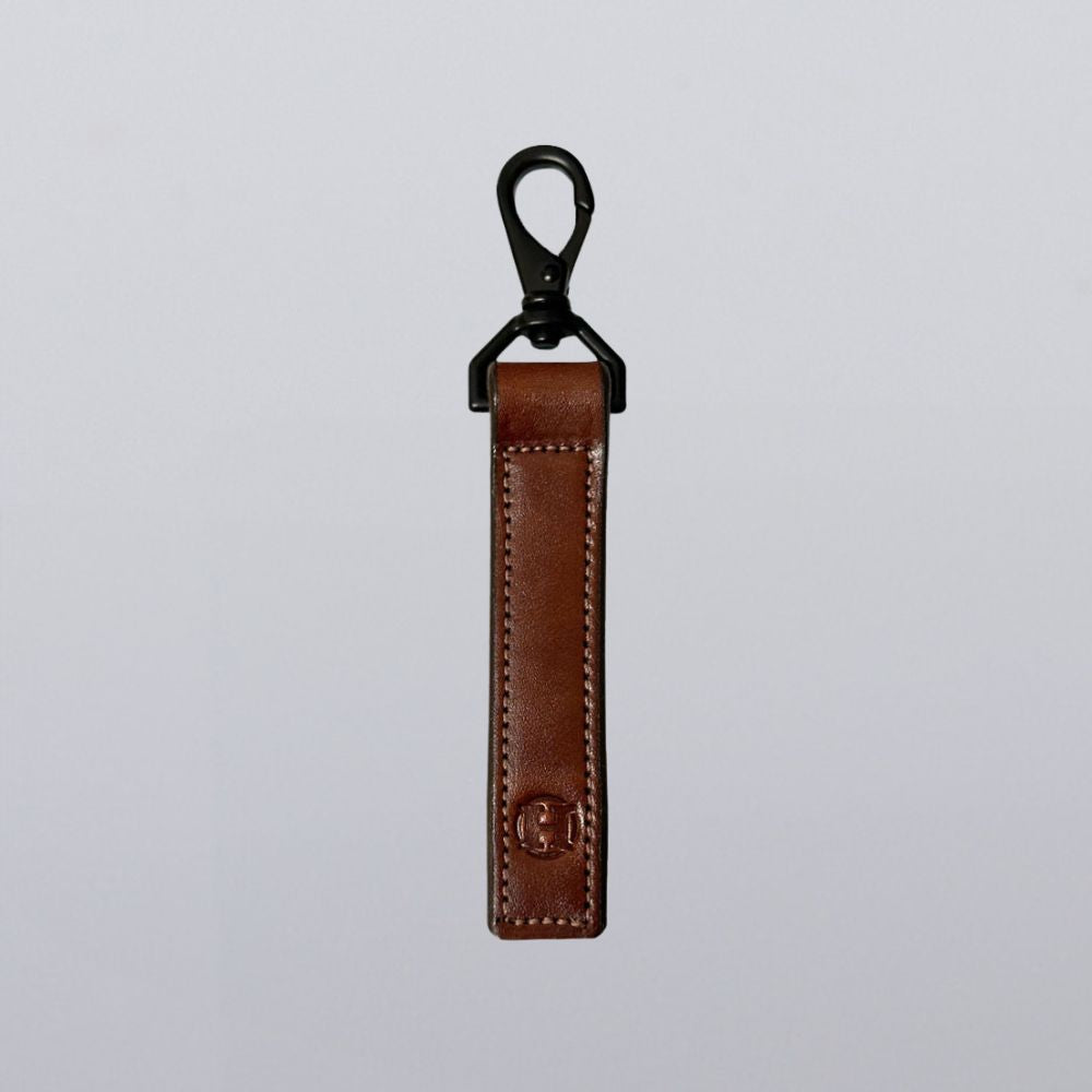 The Effortless Keychain by Hanks Belts features brown leather in a sleek rectangular design with visible stitching, an embossed logo near the bottom, and a black metal clasp, shown against a plain light gray background.