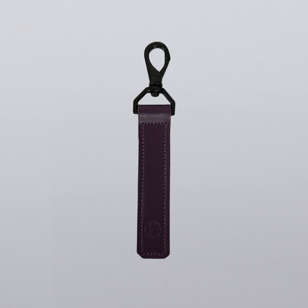 The Hanks Belts Effortless Keychain in purple leather features visible stitching, a sleek rectangular shape, and a black metal clasp, shown hanging vertically against a plain light gray background.