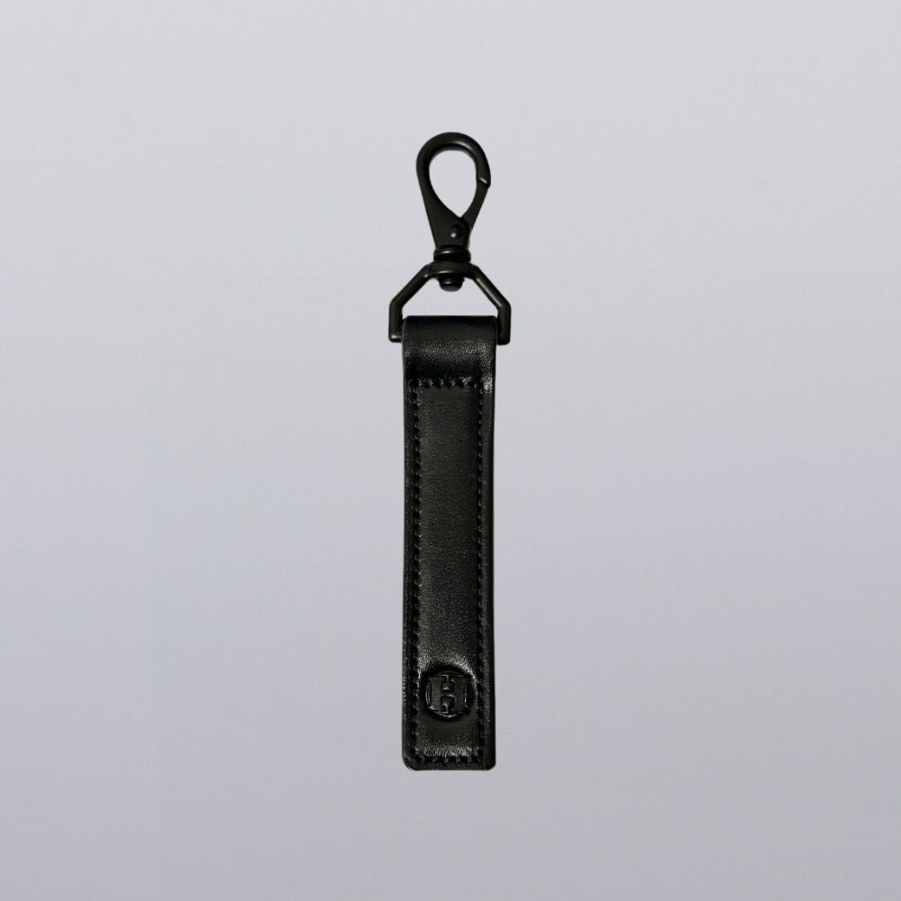 The Effortless Keychain by Hanks Belts features a sleek rectangular black leather strap with stitched edges, a metal swivel clasp, and an embossed circular logo, shown against a plain light background.
