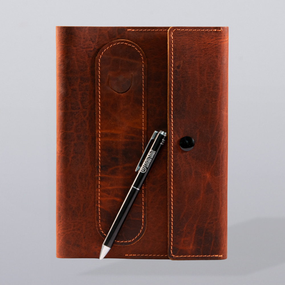 The Hanks Belts Luxury Bison Journal, featuring a refillable leather cover with flap closure and visible stitching, rests on a light surface. A black pen with white details lies diagonally—perfect for inspiring any writer.
