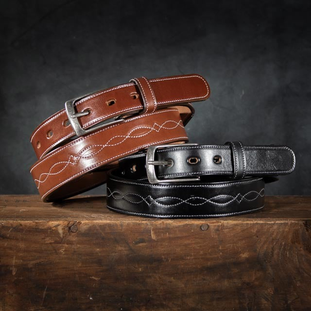 Two Teton Tapered belts by Hanks Belts, one brown and one black, crafted from vegetable-tanned leather with white infinity stitching and silver buckles, are crossed on a rustic wooden surface against a dark background.