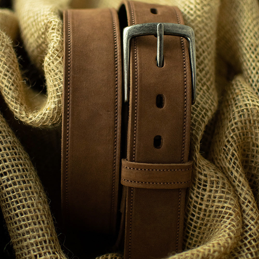The Suede - 1.5" by Hanks Belts, a USA-made brown leather belt with a silver buckle, stands upright on textured burlap, showcasing its smooth finish and detailed stitching.