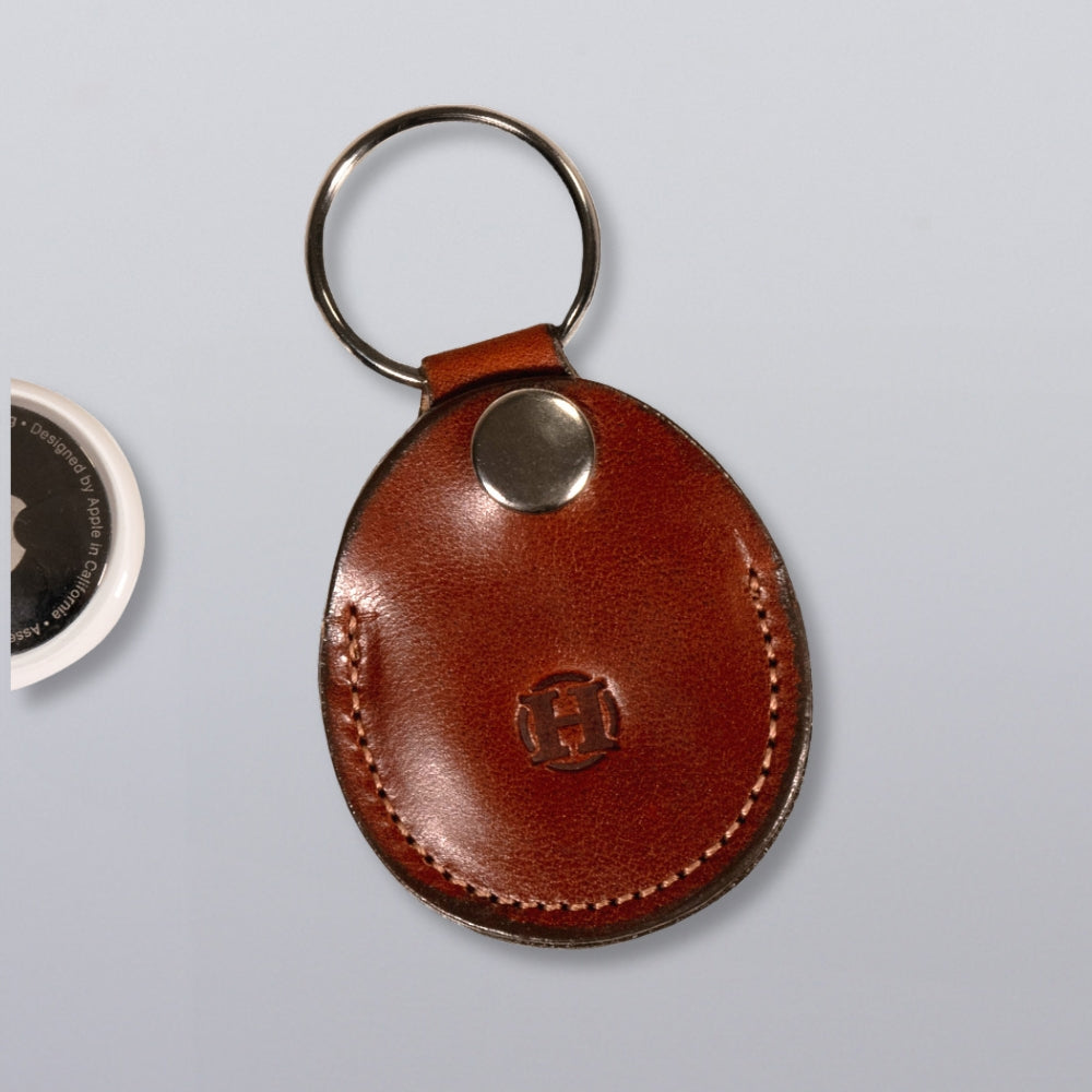 The Hanks Belts Circular Tag Holder is a brown full-grain leather keychain with visible stitching, a metal snap button, and keyring—ideal as an AirTag leather holder—shown on a light gray surface.