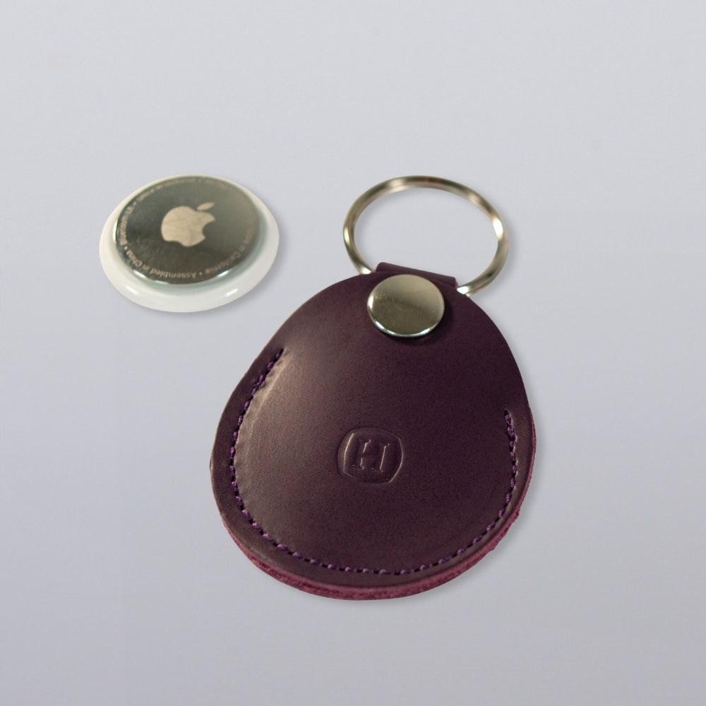 A Hanks Belts Circular Tag Holder in purple full-grain leather with a metal snap and keyring, shown with a round tag on a light gray background.