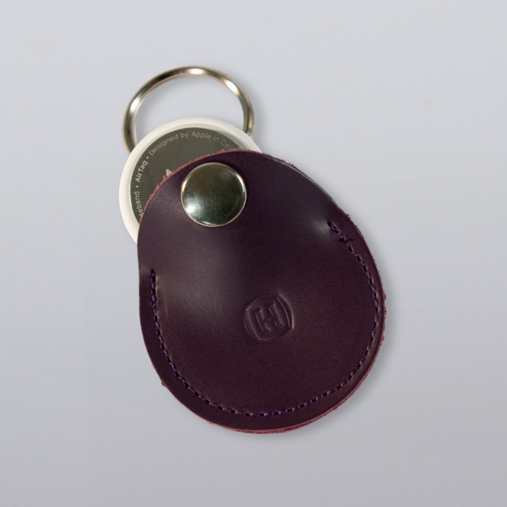 The Hanks Belts Circular Tag Holder is a purple full-grain leather keychain with a round metal button, shown securing an Apple AirTag on a plain light gray surface.