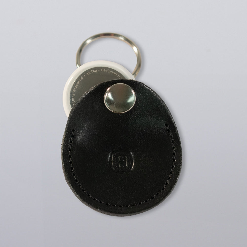 The Hanks Belts Circular Tag Holder is a black full-grain leather keychain with visible stitching, a snap button, and a white circular tag, serving as an elegant AirTag holder attached to a silver keyring on a light background.