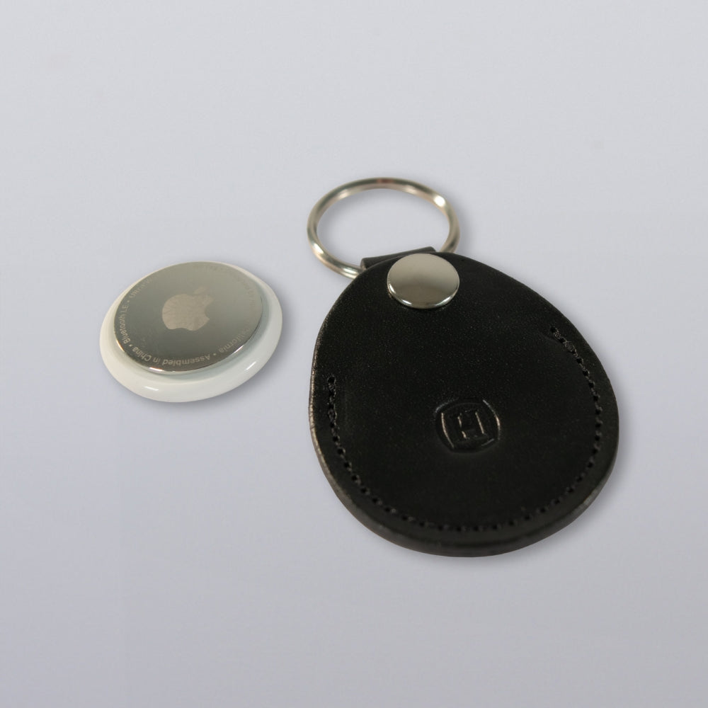 A Hanks Belts Circular Tag Holder in black full-grain leather with a metal ring rests beside a round white Apple AirTag on a plain light gray surface.