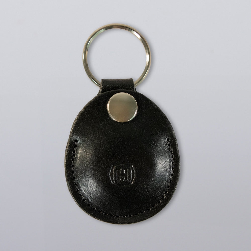 The Hanks Belts Circular Tag Holder is a black full-grain leather keychain with a round silver ring and metallic button, featuring an embossed logo on the front. It is shown against a plain, light gray background.