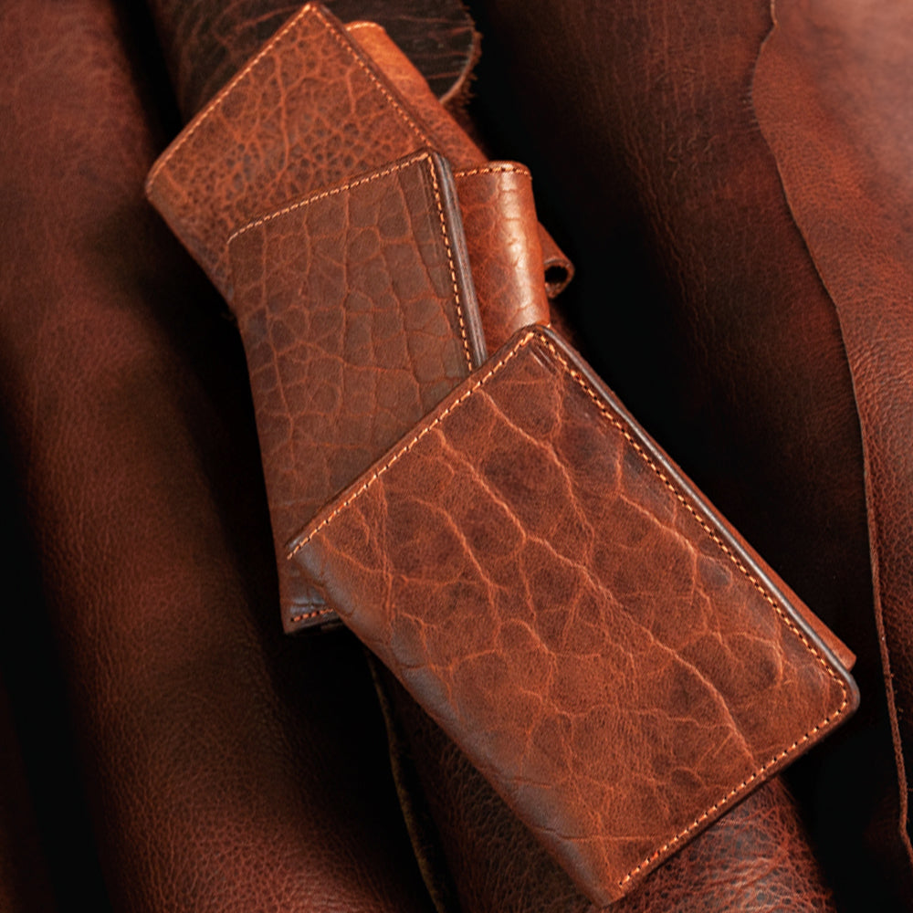 Three Hanks Belts Erie Premium Bison Trifold wallets with visible grain rest on matching brown leather. Each wallet features clean stitching, highlighting rugged texture and exceptional craftsmanship.