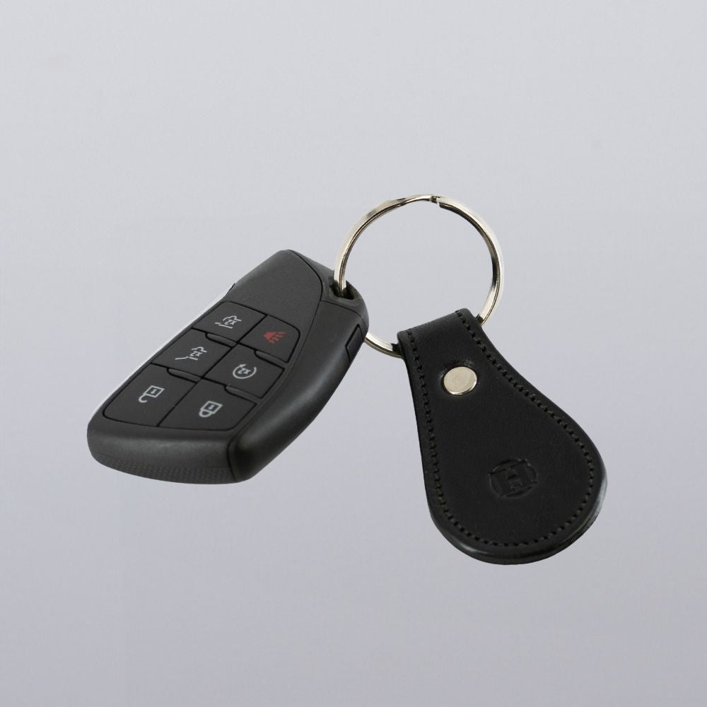 A black car key fob with multiple buttons is attached to a round metal keyring, which also holds the Hanks Belts Bell-Shaped Leather Keychain made from full grain leather. The background is plain light gray.