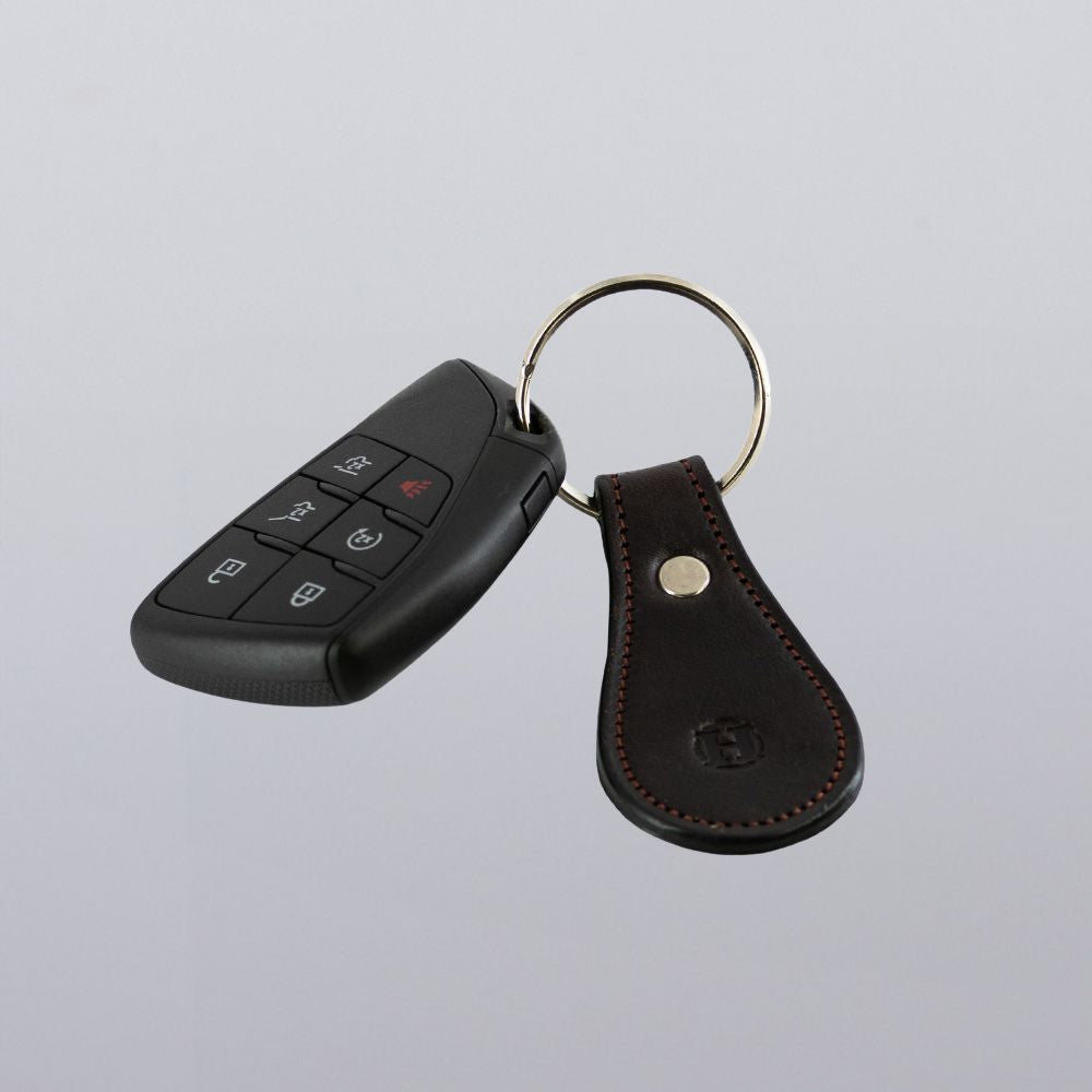 A Hanks Belts Bell-Shaped Leather Keychain, made from full grain leather with visible stitching, is attached to a black car key fob and a round metal key ring, displayed on a plain light gray background.