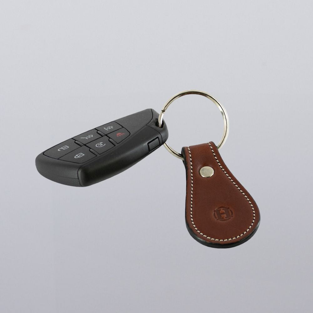 A black car key fob with four buttons is attached to a round metal keyring and a Hanks Belts Bell-Shaped Leather Keychain made from full grain leather with white stitching, all on a light gray background.