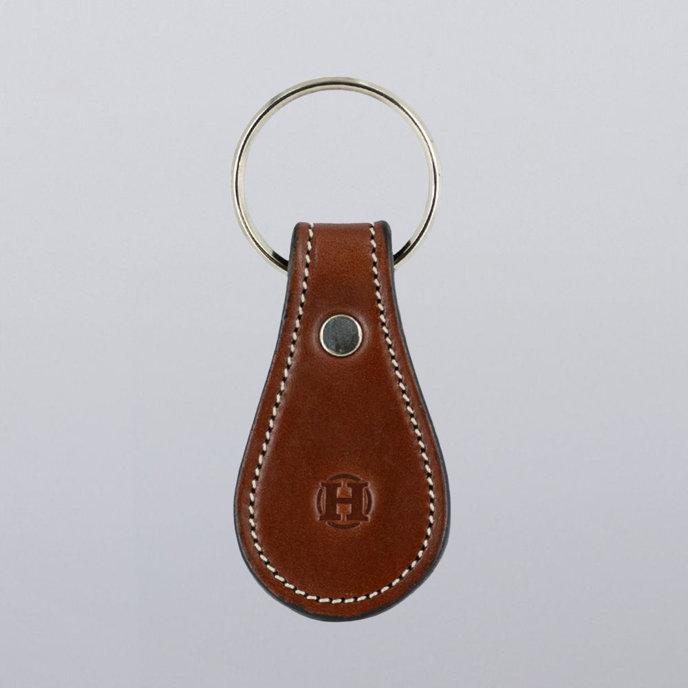 The Hanks Belts Bell-Shaped Leather Keychain is crafted from full grain leather with white stitching, a silver metal keyring, and an embossed logo. Shown against a plain light gray background.