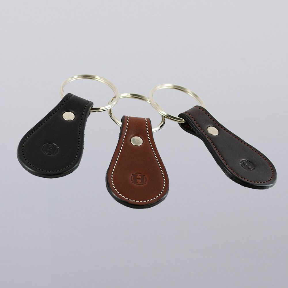 Three Hanks Belts Bell-Shaped Leather Keychains—black, brown, and dark brown—are shown side by side on a light gray surface. Each features full grain leather, metal rings, detailed stitching, and an embossed logo.