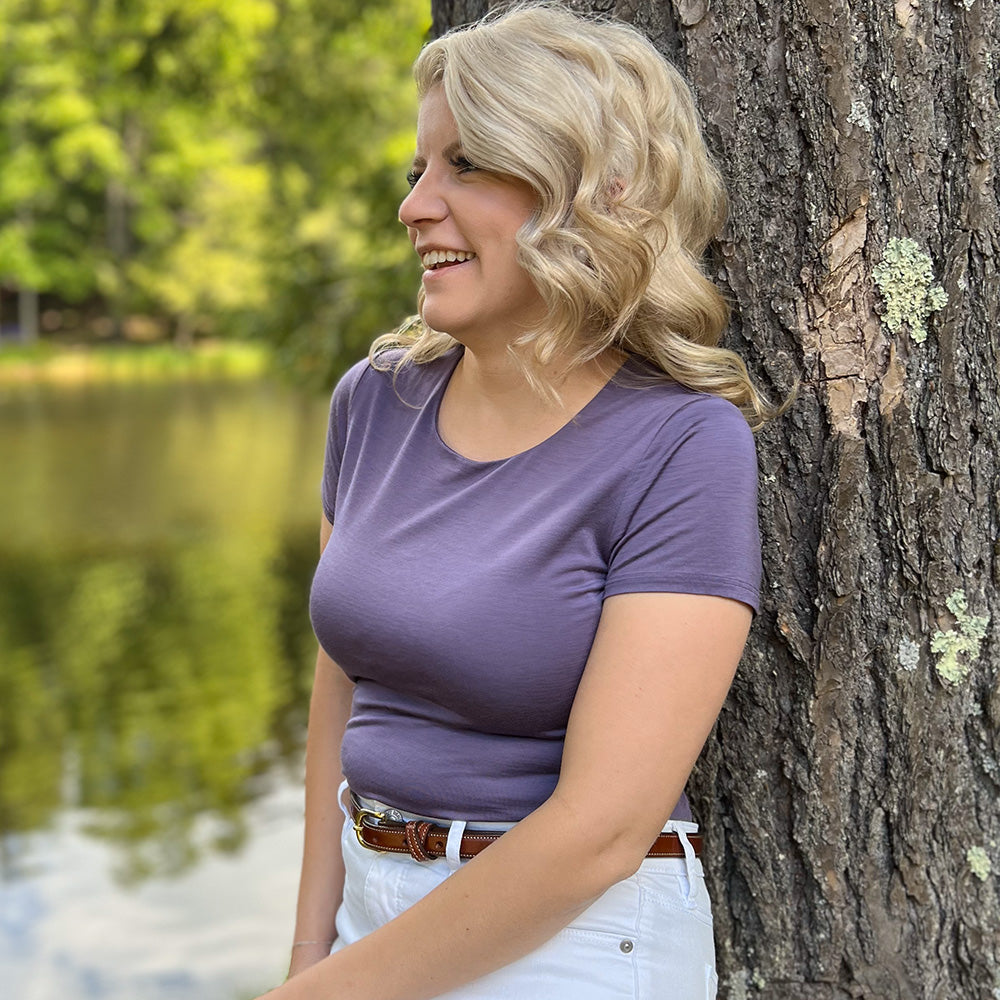A smiling woman with wavy blonde hair, dressed in a purple t-shirt, white pants, and The Dixie women's belt by Hanks Belts, leans against a tree by a pond with green trees in the background.