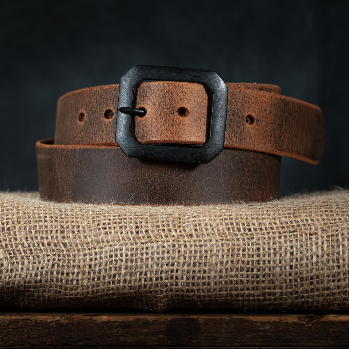The Relic Retro Style by Hanks Belts, a coiled brown Water Buffalo leather belt with a square black buckle, is displayed on burlap fabric against a dark backdrop.