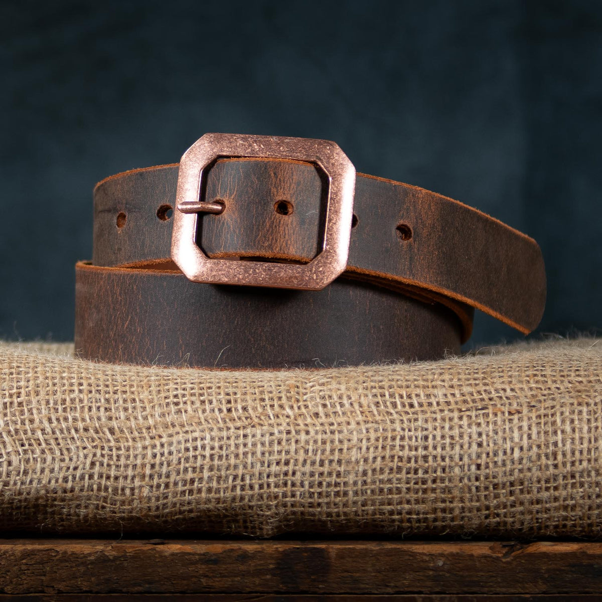 The Relic Retro Style by Hanks Belts—a Water Buffalo leather belt with a copper-tone square buckle—is coiled on burlap fabric against a dark background.
