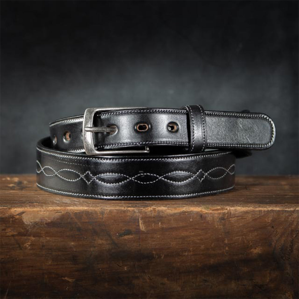 The Teton Tapered by Hanks Belts, in black vegetable tanned leather with a silver buckle and white infinity stitching, is coiled on a rustic wooden surface against a dark background.