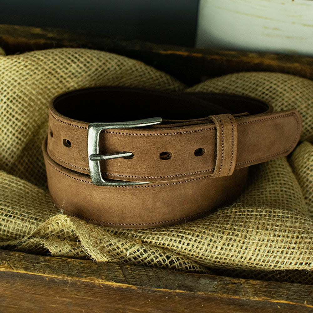 The Suede - 1.5" by Hanks Belts, a brown nubuck leather belt with a silver buckle, is coiled on textured burlap inside a wooden box.