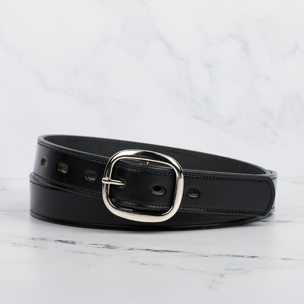 The Eloise by Hanks Belts, a women's premium black vegetable-tanned leather belt with a shiny silver center bar buckle, is coiled on a light marble surface against a white marble background.