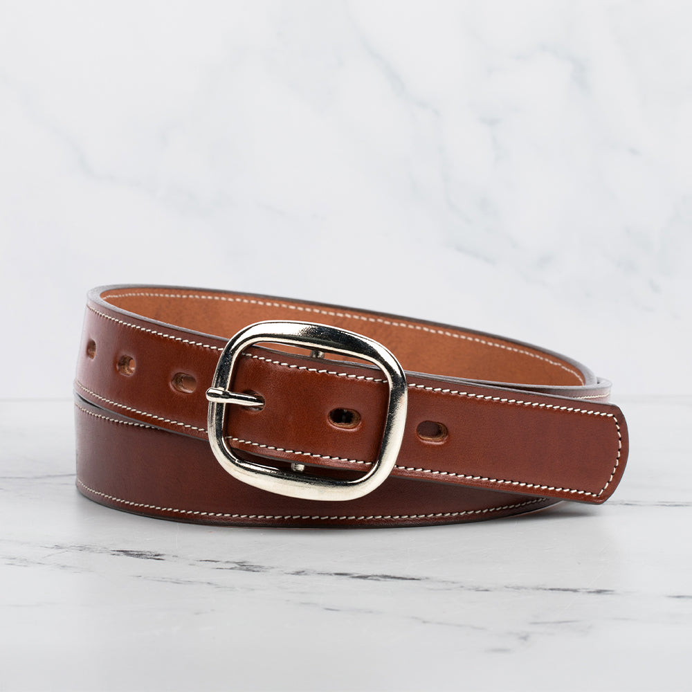 The Eloise by Hanks Belts is a women's premium brown vegetable-tanned leather belt with white stitching and a silver center bar buckle, coiled on a white marble surface against a light gray marbled background.