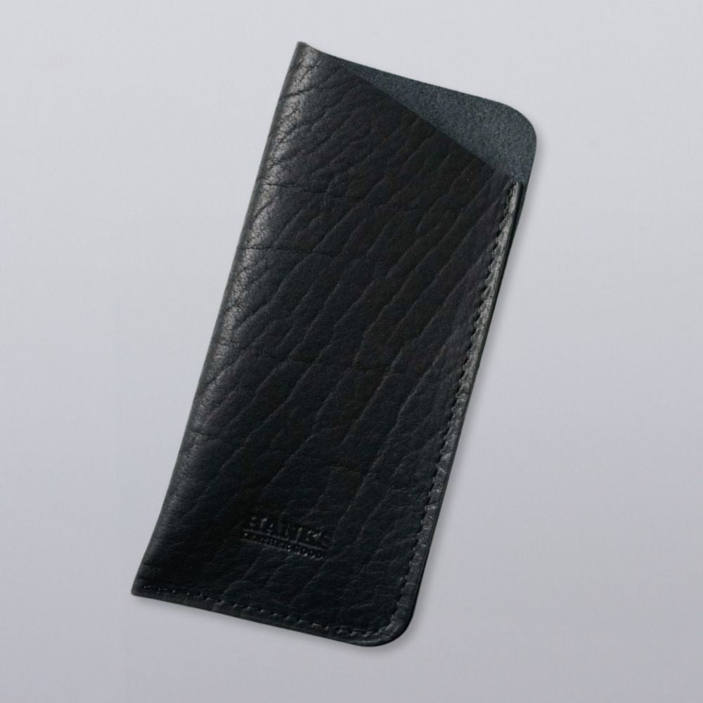 A black, textured Bison Leather Eyeglass Case by Hanks Belts features visible stitching along the right edge, displayed on a light gray background. "Hanks Belts" is embossed near the bottom. Made in the USA.