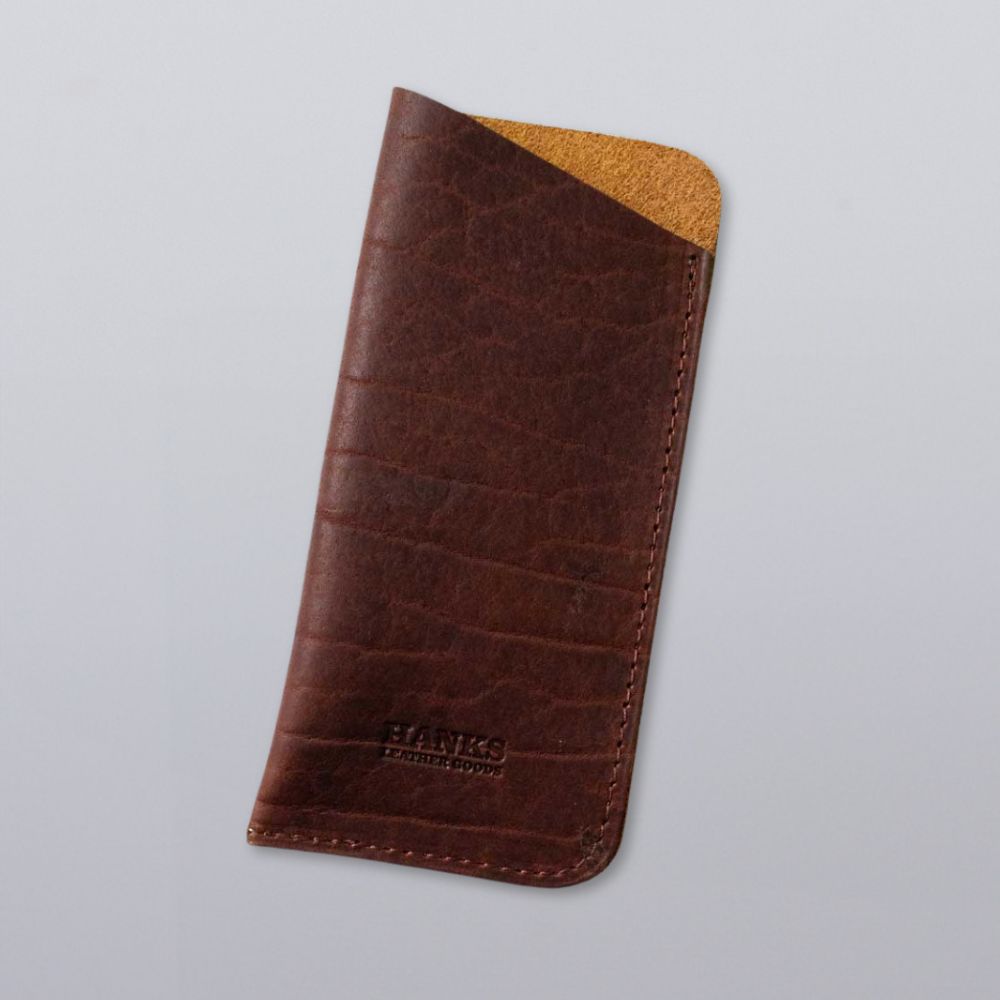 A brown Bison Leather Eyeglass Case by Hanks Belts, with tan suede interior and edge stitching, features the brand embossed on the front. Made in the USA, it’s shown on a plain light gray background.