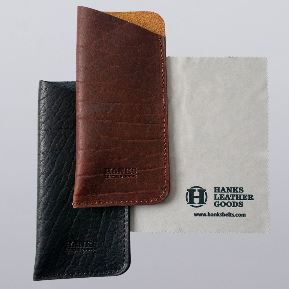 Two leather phone sleeves, one brown and one black, sit beside a gray cleaning cloth printed with "Hanks Belts" and their website—an ideal match for your Bison Leather Eyeglass Case.