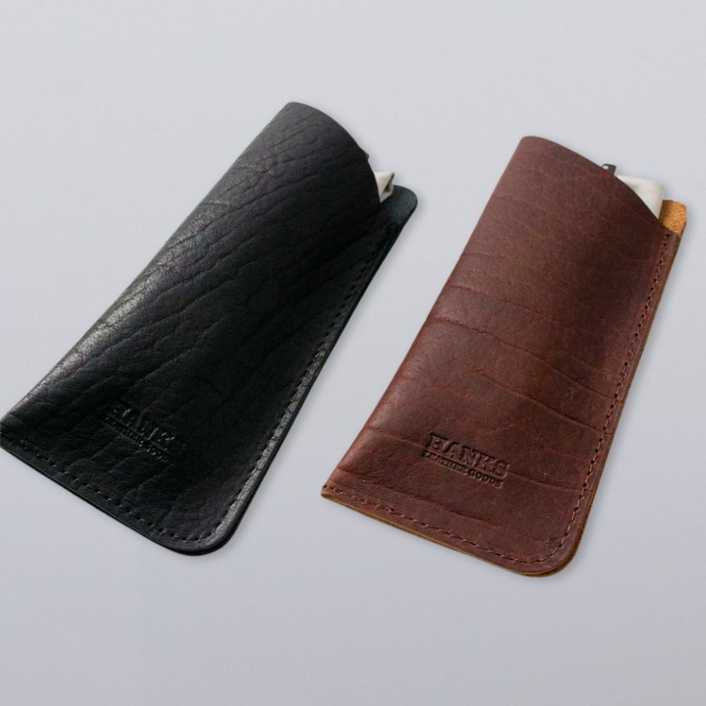Two Hanks Belts Bison Leather Eyeglass Cases—one black, one brown—are shown side by side on a light gray background. Both cases display embossed branding and contain a partially visible white cloth inside.