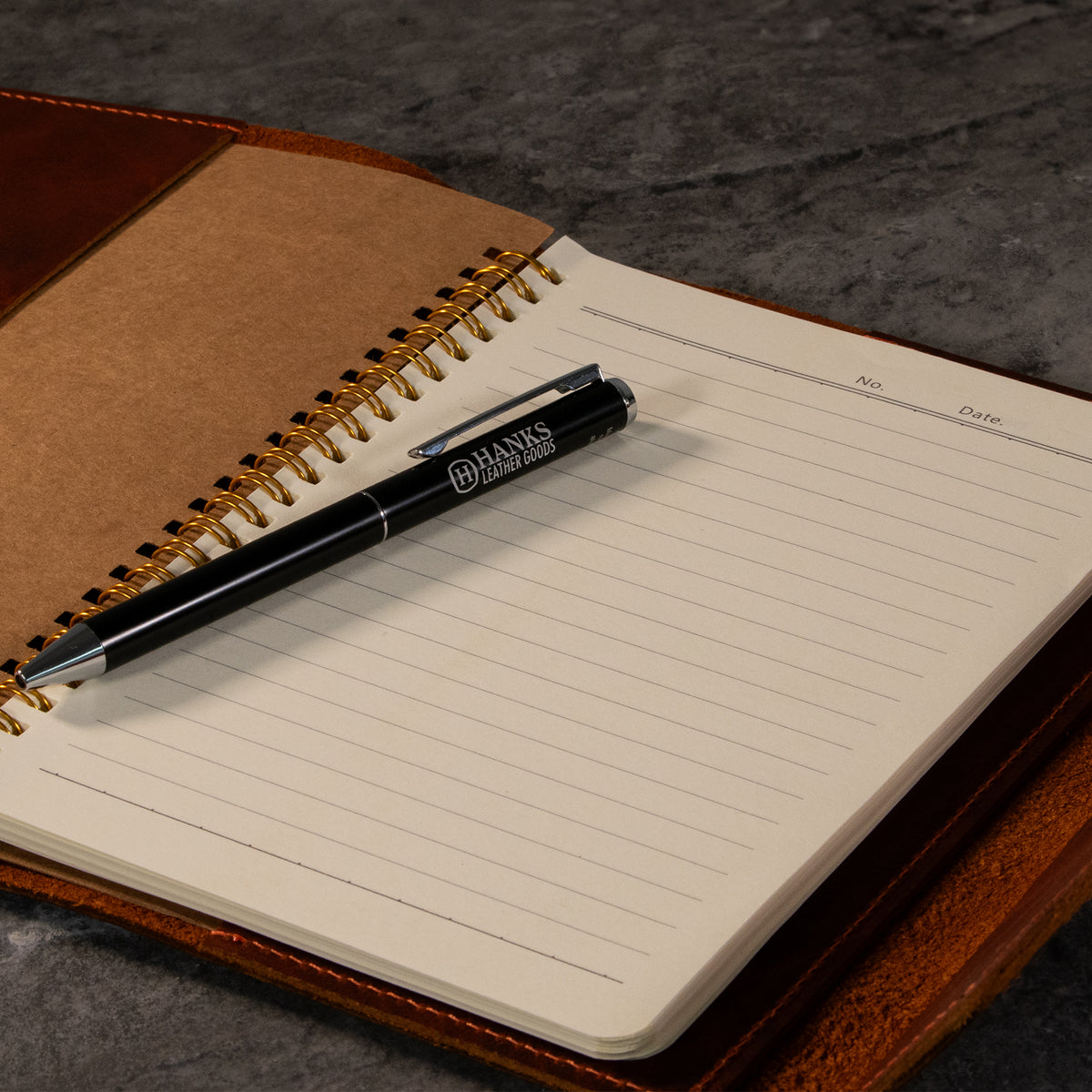 A black pen rests diagonally on the open, lined pages of the Hanks Belts Luxury Bison Journal, featuring a brown leather cover and set on a dark stone surface.