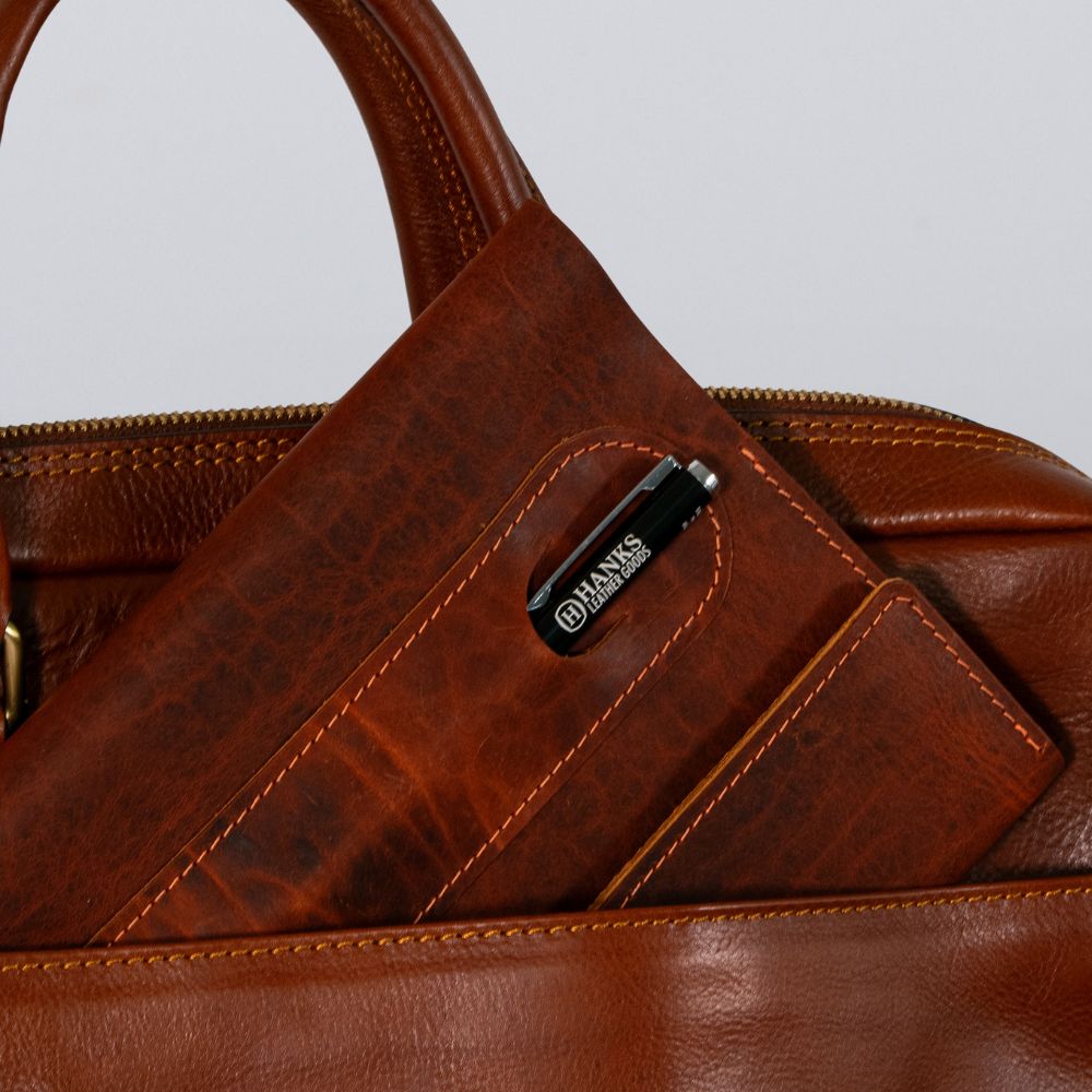 A close-up of a brown leather bag with a Luxury Bison Journal by Hanks Belts and a black pen, both tucked into the front pocket.