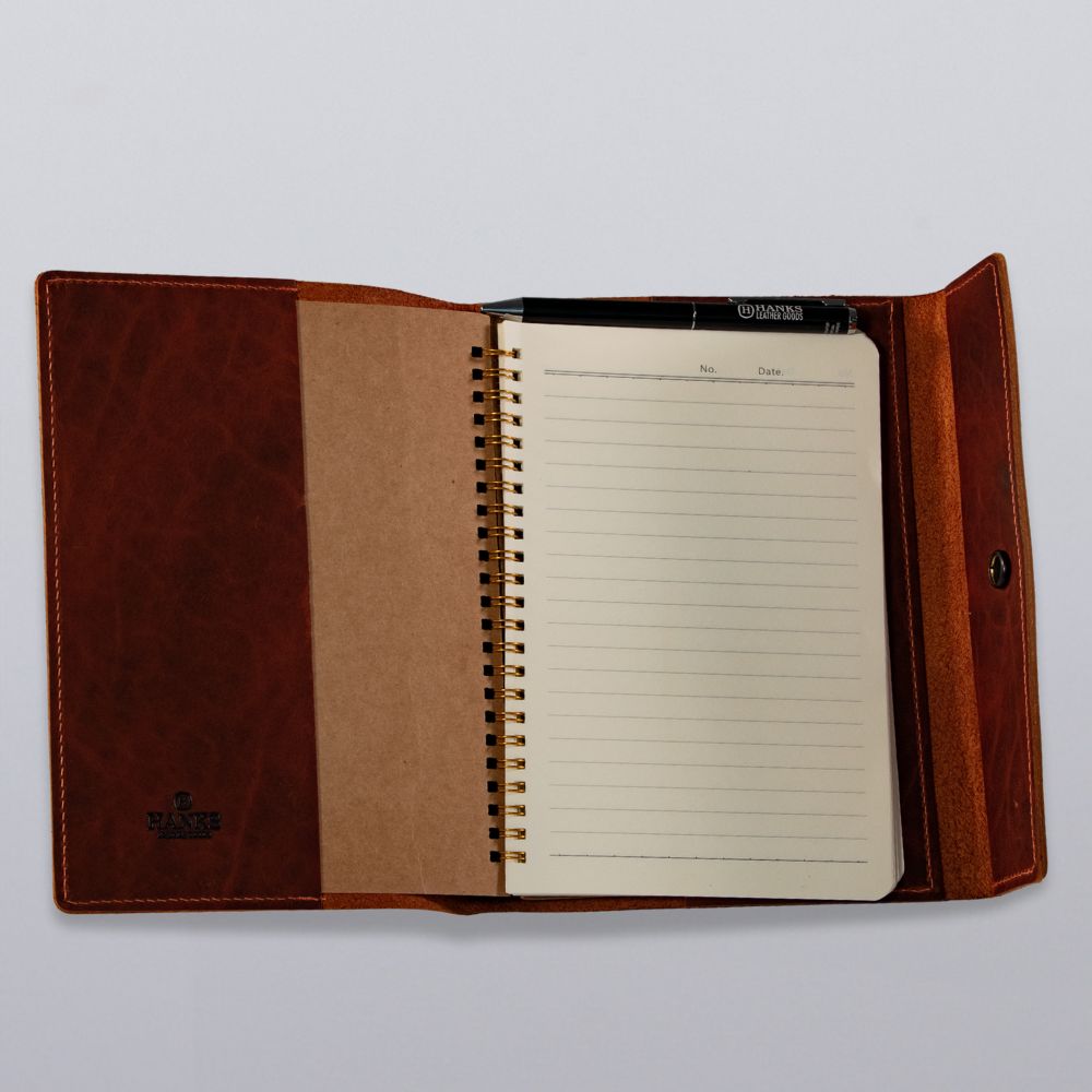 The Hanks Belts Luxury Bison Journal is an open brown leather cover with a spiral-bound lined notepad and black pen inside, showcased on a plain white surface.