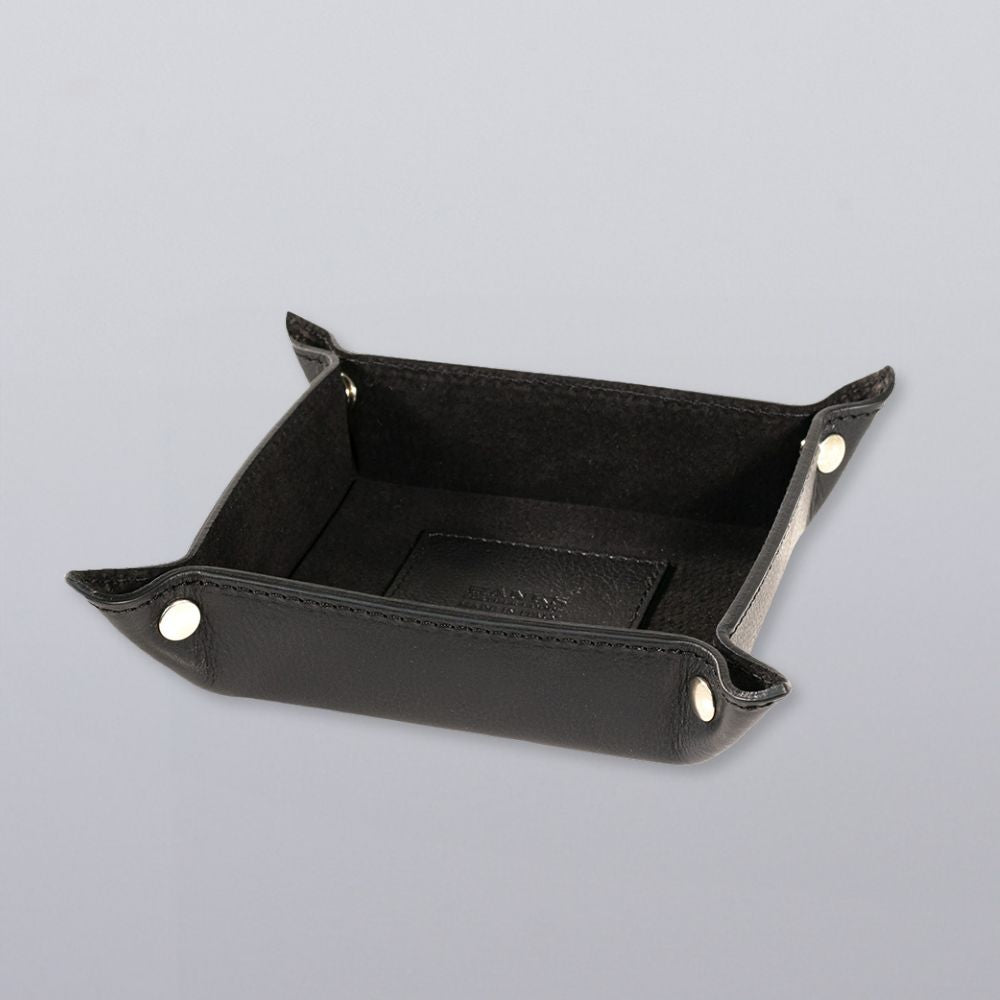 The Hanks Belts Italian Compact Valet Tray, crafted from black Italian leather with snap buttons and raised sides, offers a sleek, structured design—perfect as a travel catchall. Shown here on a light gray surface.