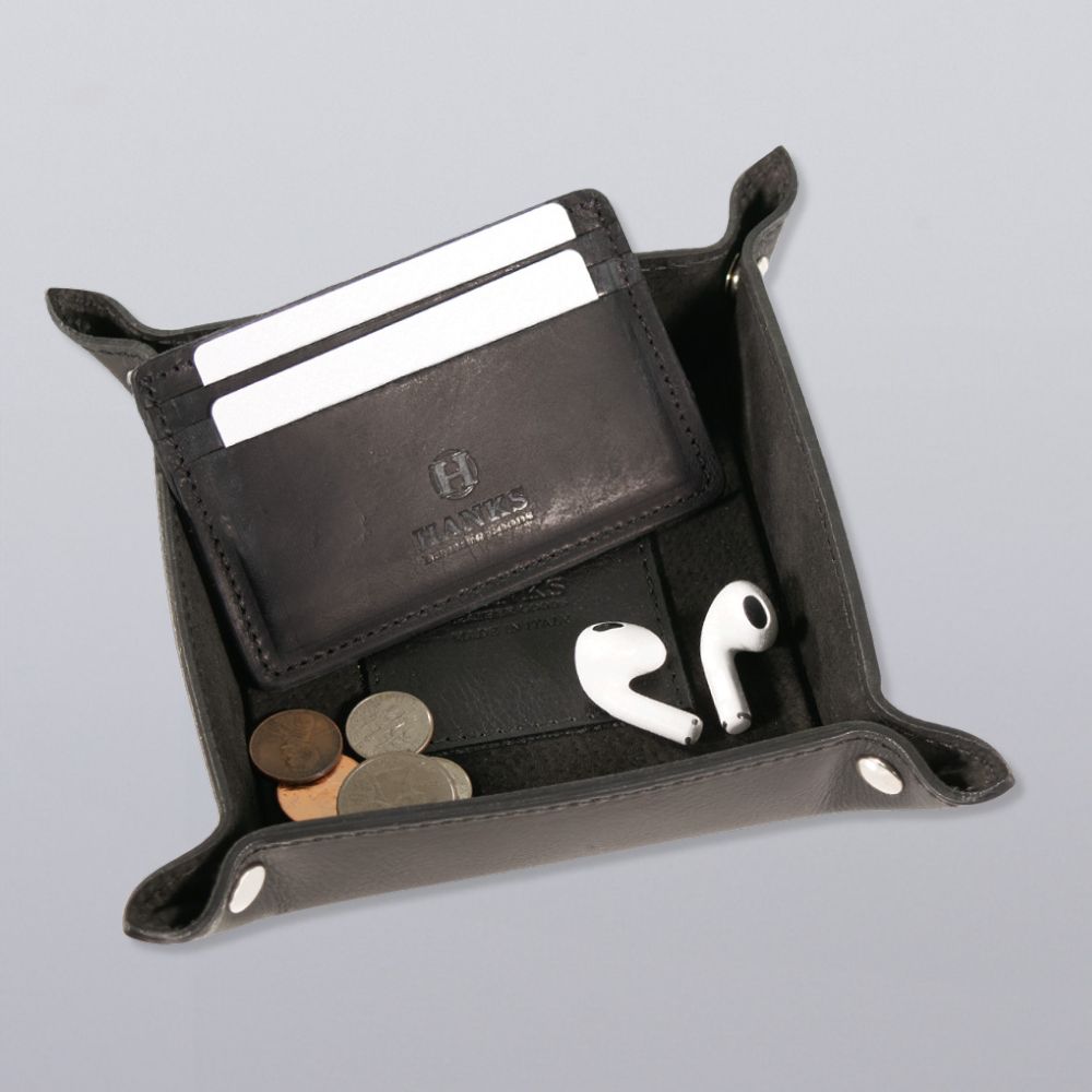 The Hanks Belts Italian Compact Valet Tray holds a wallet, cards, coins, and white wireless earbuds on a light gray surface—an elegant catchall for organizing your essentials while traveling.