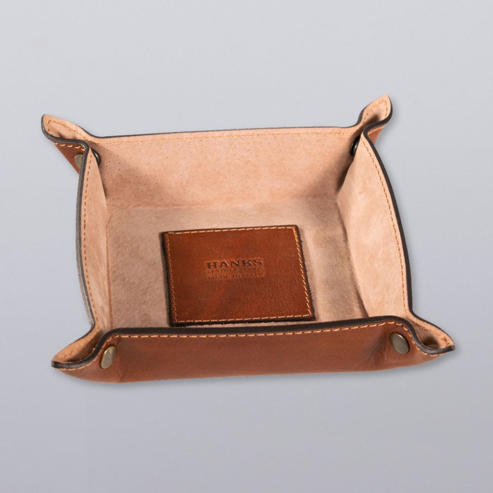 The Hanks Belts Italian Compact Valet Tray features stitched edges, metal rivets, and an embossed square leather patch inside. This leather travel catchall is shown resting on a light gray surface.