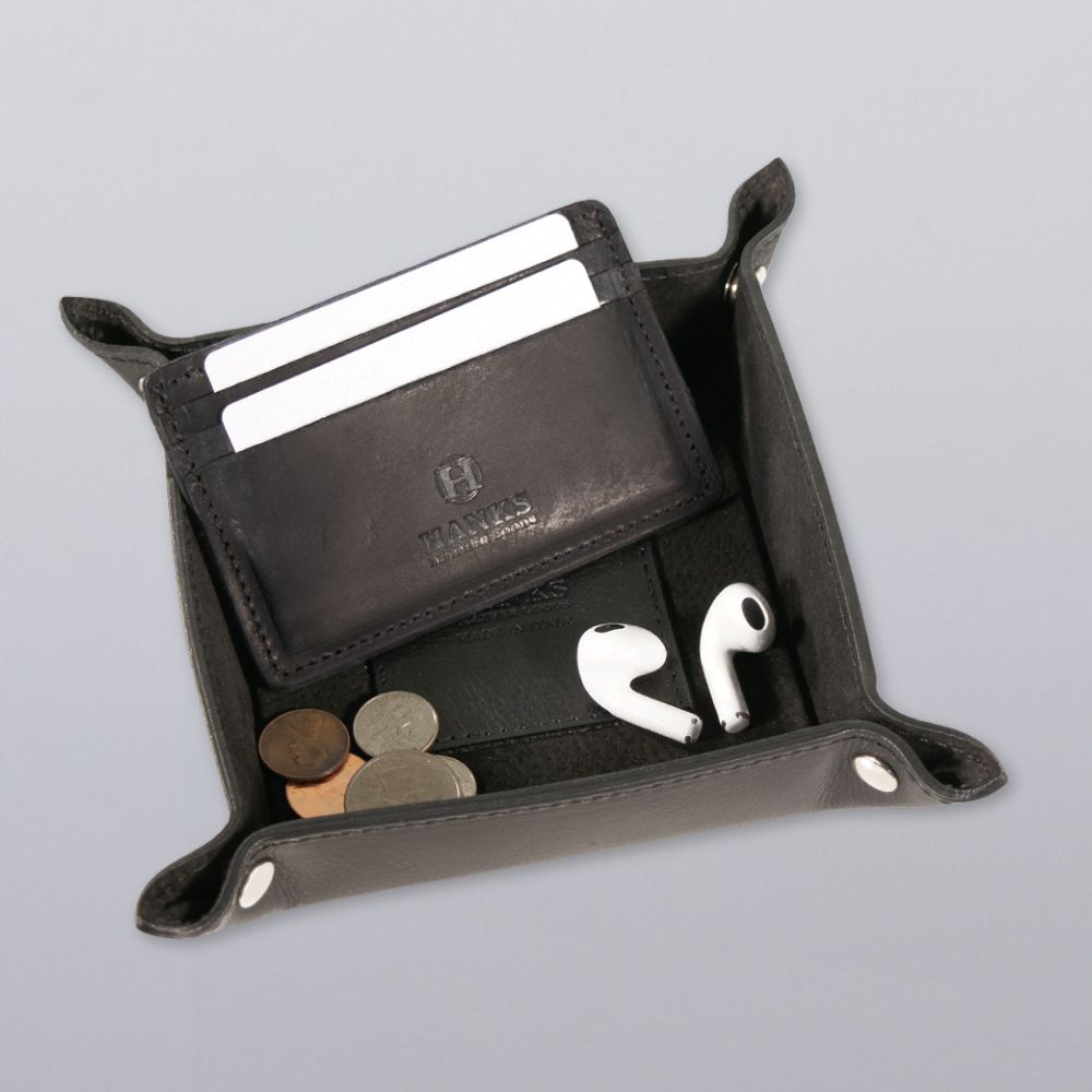 The Hanks Belts Italian Large Valet Tray in black displays a slim cardholder with three cards, several coins, and white wireless earbuds arranged neatly on a light gray surface.