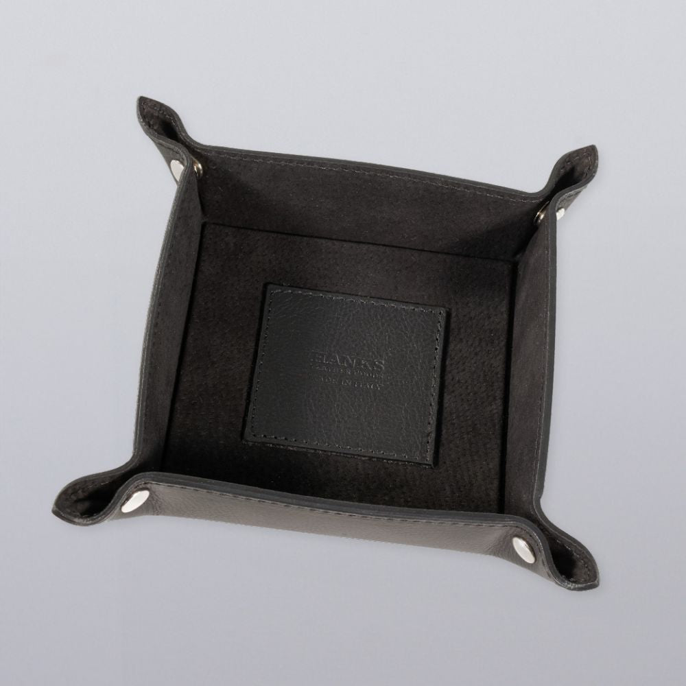 The Hanks Belts Italian Large Valet Tray is a square, dark gray leather tray with raised sides and corner snap buttons, shown from above on a light gray surface. The interior features a stitched patch with embossed text.