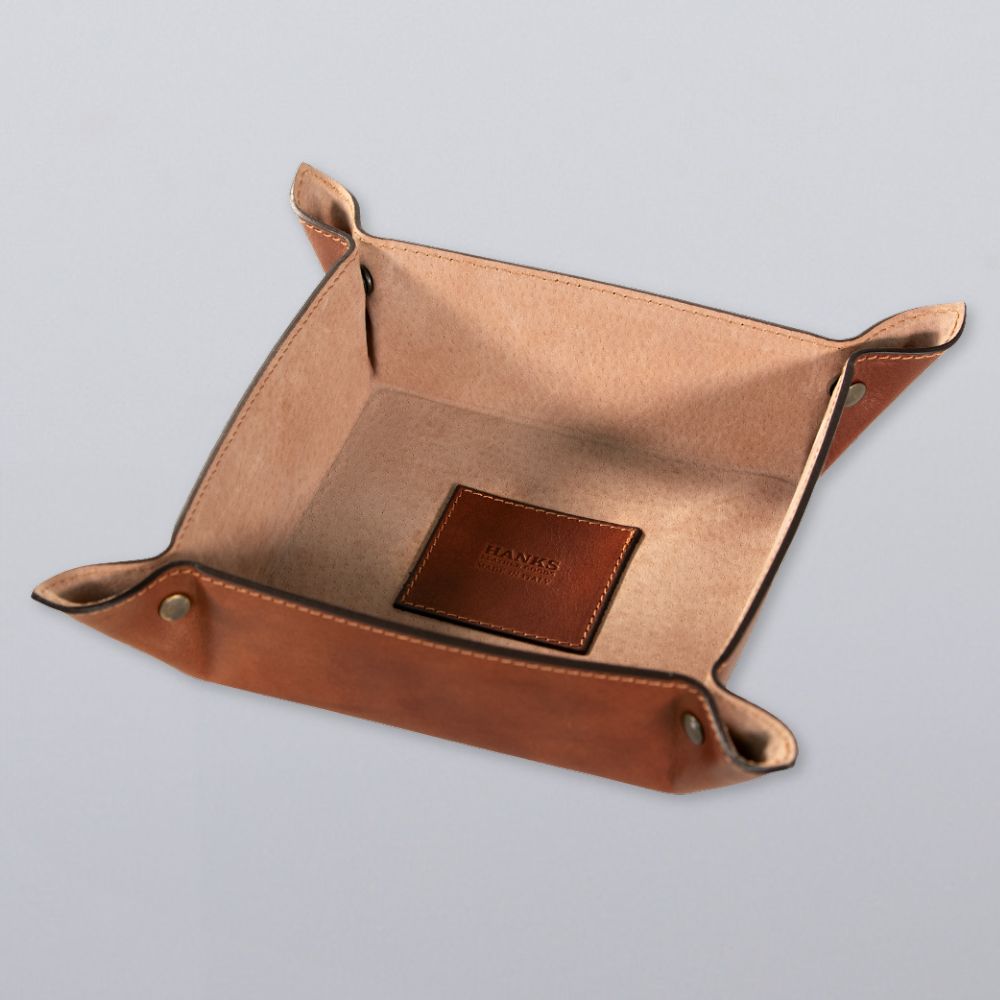 The Hanks Belts Italian Large Valet Tray, made of square Italian leather with raised sides and metal snaps at each corner, rests on a light gray surface. A brown leather patch with embossed text inside adds style and function for travel.