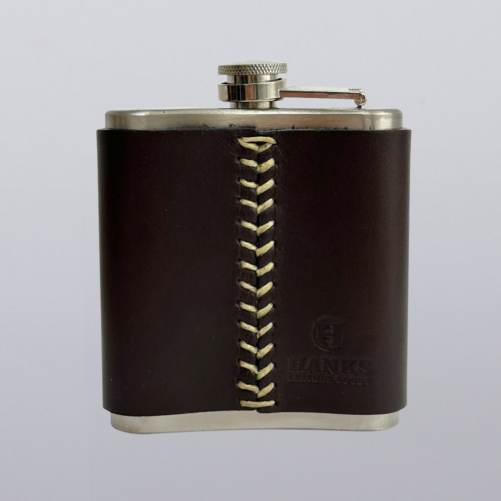 The Hanks Belts Leather Covered Flask is a USA-made stainless steel flask, wrapped in dark brown leather with light center stitching, a secure screw-top lid, and embossed “Hanks Leather Co.” on the lower right.