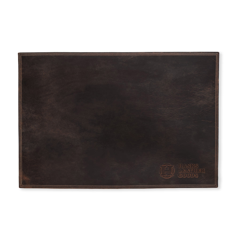A rectangular dark leather desk pad, crafted in the USA from North American Bison leather, proudly displays the embossed "Hanks Belts" logo in the bottom right corner. It showcases visible stitching along the edges, providing a classic, polished look that enhances any desk setup. Known as the Desk & Mouse Pad Set.