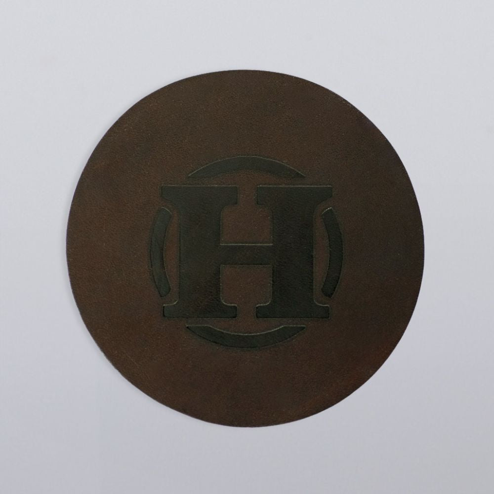 A round, dark brown Hanks Belts leather coaster from the "Round Leather Coasters - Set of 4," embossed with a bold “H” and two curved lines, displayed on a plain light gray background.