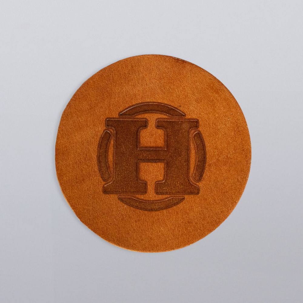 A set of four premium round leather coasters by Hanks Belts, each featuring an embossed "H" and a decorative border, displayed on a light gray background. Part of our USA Made Leather Coasters collection.