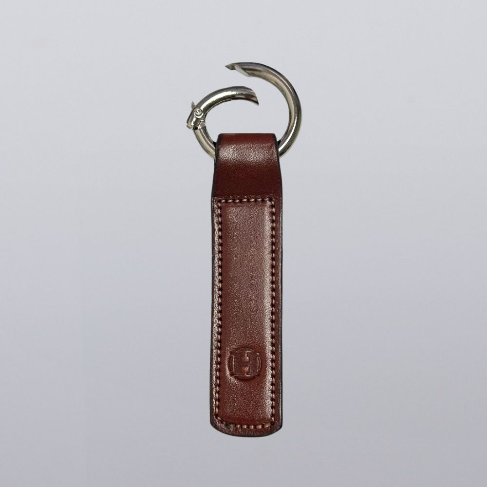 The Superior Keychain from Hanks Belts is a brown leather rectangle with visible stitching, embossed logo near the bottom, and a metal open gate keyring at the top, displayed on a plain light gray background.