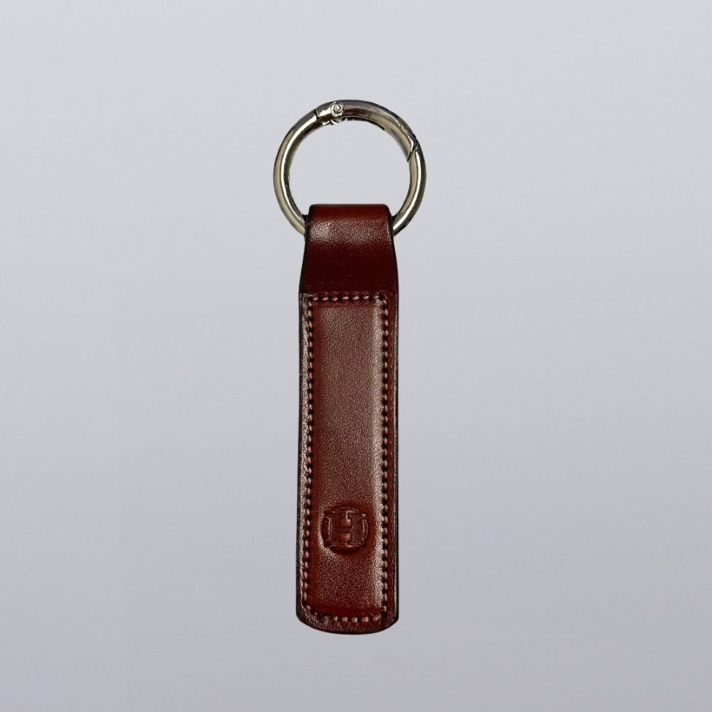 The Superior Keychain by Hanks Belts is a brown leather rectangle with visible stitching, an open gate keyring, and a subtle embossed logo near the bottom, displayed on a plain light gray background.