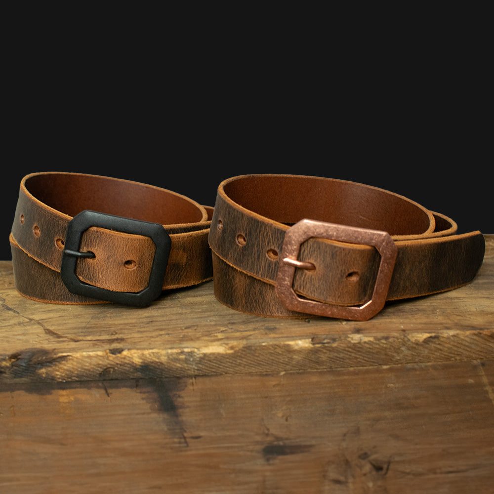 Two brown "The Relic Retro Style" Water Buffalo leather belts by Hanks Belts, with square black and bronze buckles, are coiled on rustic wood against a dark background—American-made classics known for durability and timeless style.