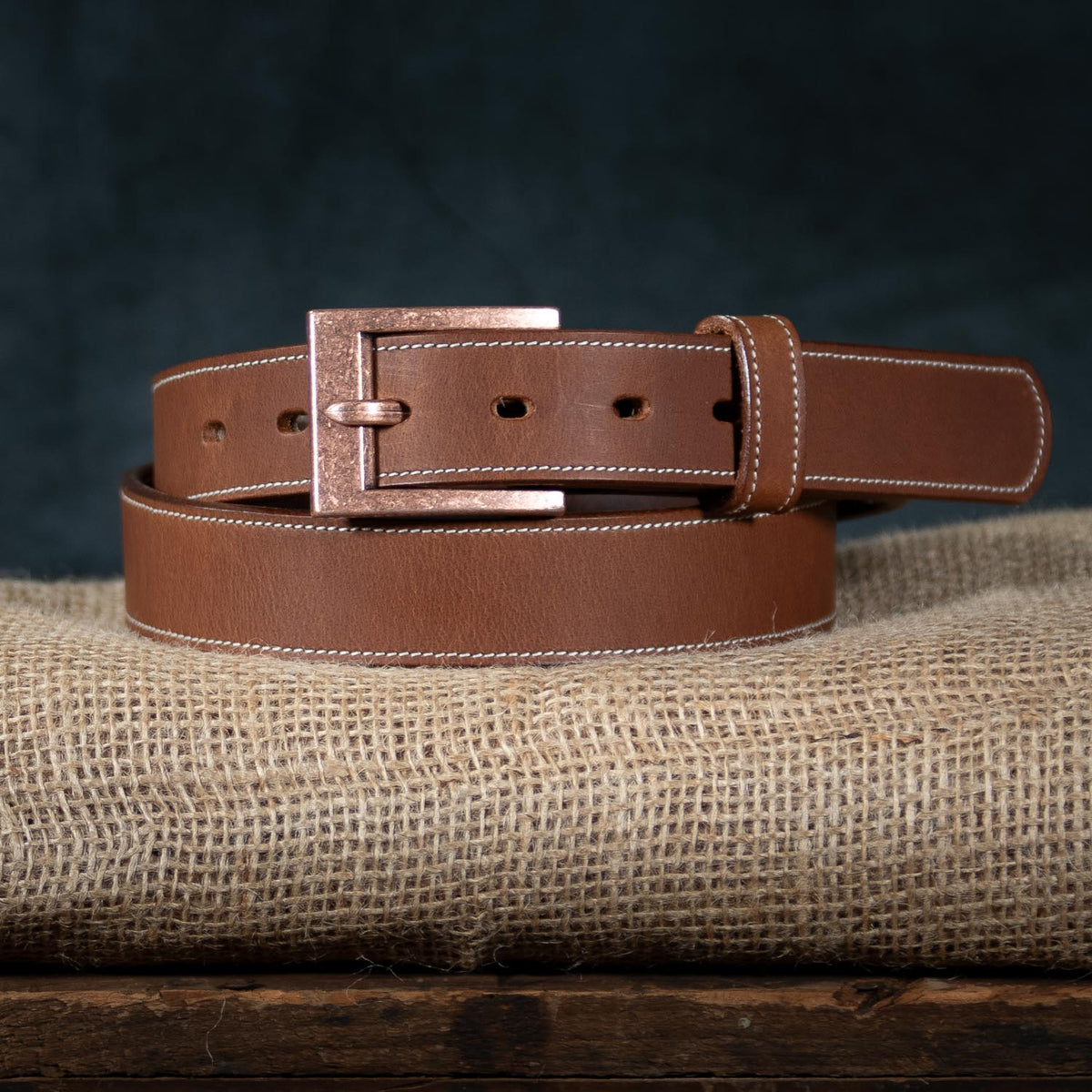 The Heritage Bison by Hanks Belts, featuring white stitching and a copper buckle, is coiled on burlap against a dark backdrop—highlighting American craftsmanship and rugged style.