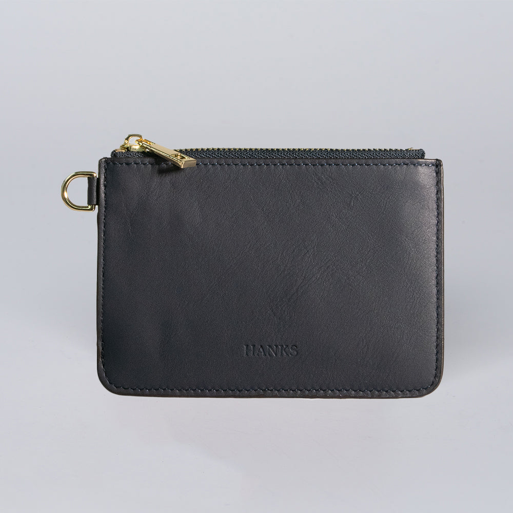 The Livia Mini Wristlet by Hanks Belts is a handcrafted black leather zippered pouch with a gold zipper, left-side metal ring, and "HANKS" embossed on the front, shown against a plain light background.