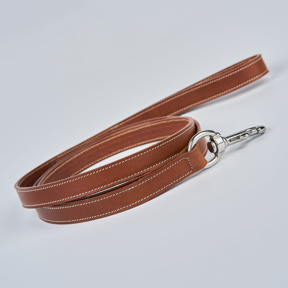 The Ollie Bison Leash by Hanks Belts, crafted from premium bison leather with white stitching and a silver metal clasp and ring, is coiled neatly on a light gray surface.