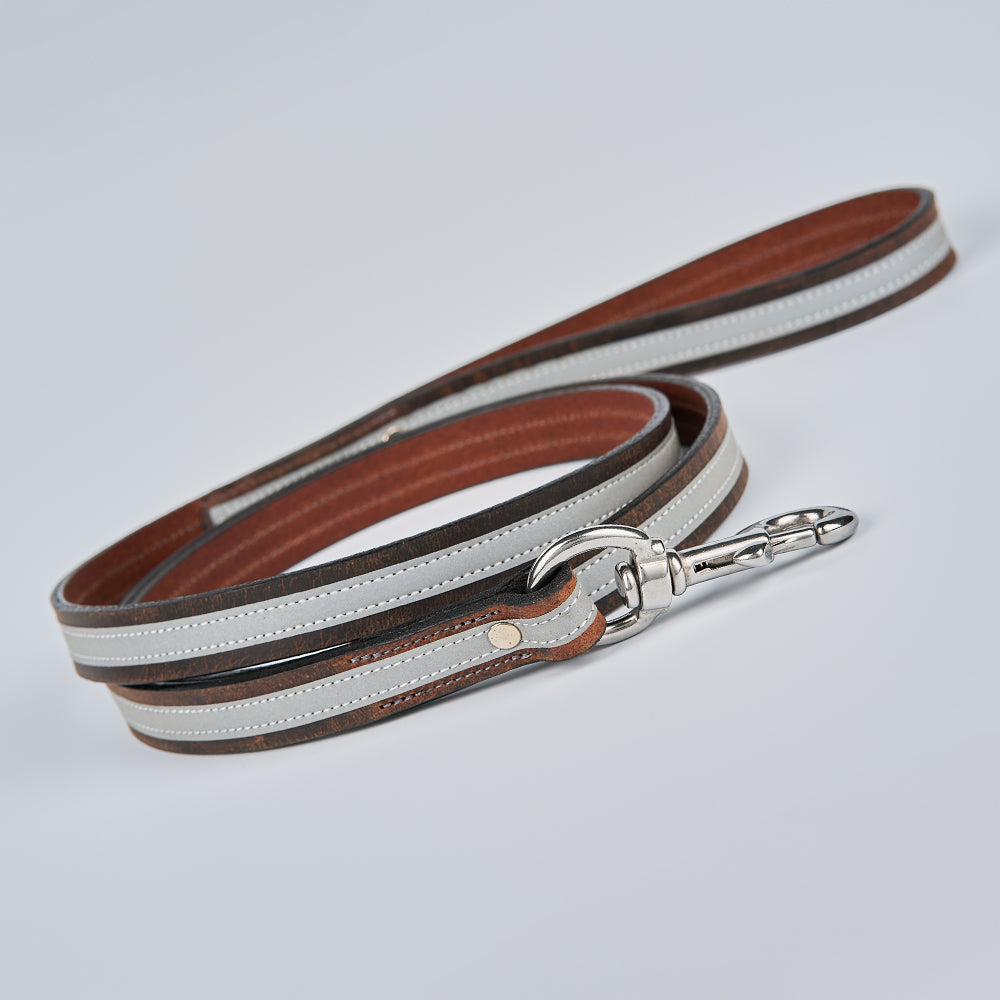 The Remi Reflective Leash by Hanks Belts, crafted from brown and gray bison leather with white stitching and a silver metal clasp, is coiled neatly on a light gray surface—ideal for evening dog walks.